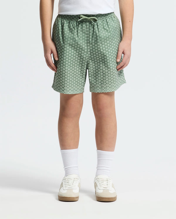 A person in a white shirt wears Psycho Bunny's KIDS BENDIGO PRINTED SWIM TRUNK, featuring a subtle bunny pattern, an elastic waistband, and a drawstring. They pair it with white socks and sneakers against a plain white background.