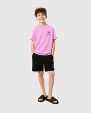 A young boy with curly hair is smiling and wearing a pink t-shirt with a small bunny graphic, Psycho Bunny KIDS PARKER HYDROCHROMIC SWIM TRUNK - B0W646C200 made of quick-dry material, and black slides. His hands are in his pockets, and he stands against a plain gray background.