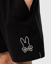 Close-up of a person wearing Psycho Bunny KIDS PARKER HYDROCHROMIC SWIM TRUNK - B0W646C200 with one hand in the pocket. The black shorts feature a graphic of a bunny head above two crossed bones in white, printed near the bottom hem. Made from quick-dry material, these stylish trunks stand out against the plain, light-colored background.