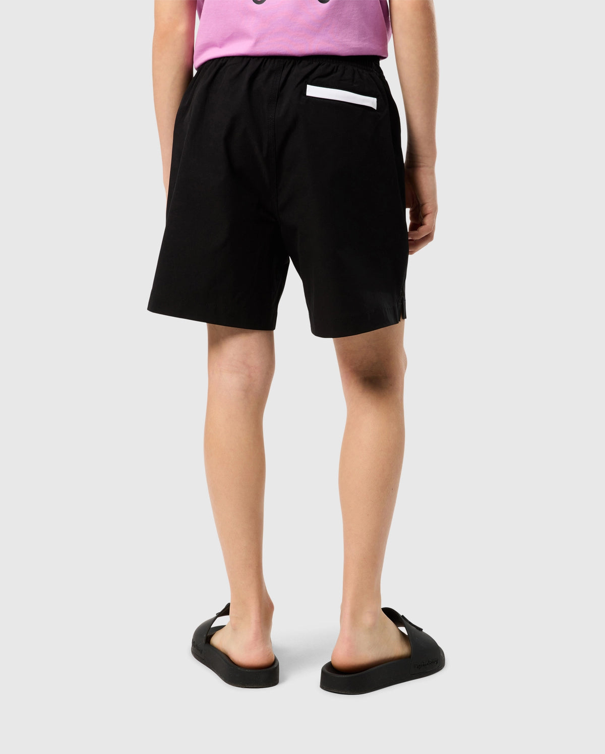A person wearing Psycho Bunny KIDS PARKER HYDROCHROMIC SWIM TRUNK - B0W646C200, a light purple top, and black sandals stands facing away from the camera. The swim trunks feature a white accent on one of the back pockets and are made of quick-dry material. The background is plain and light-colored.