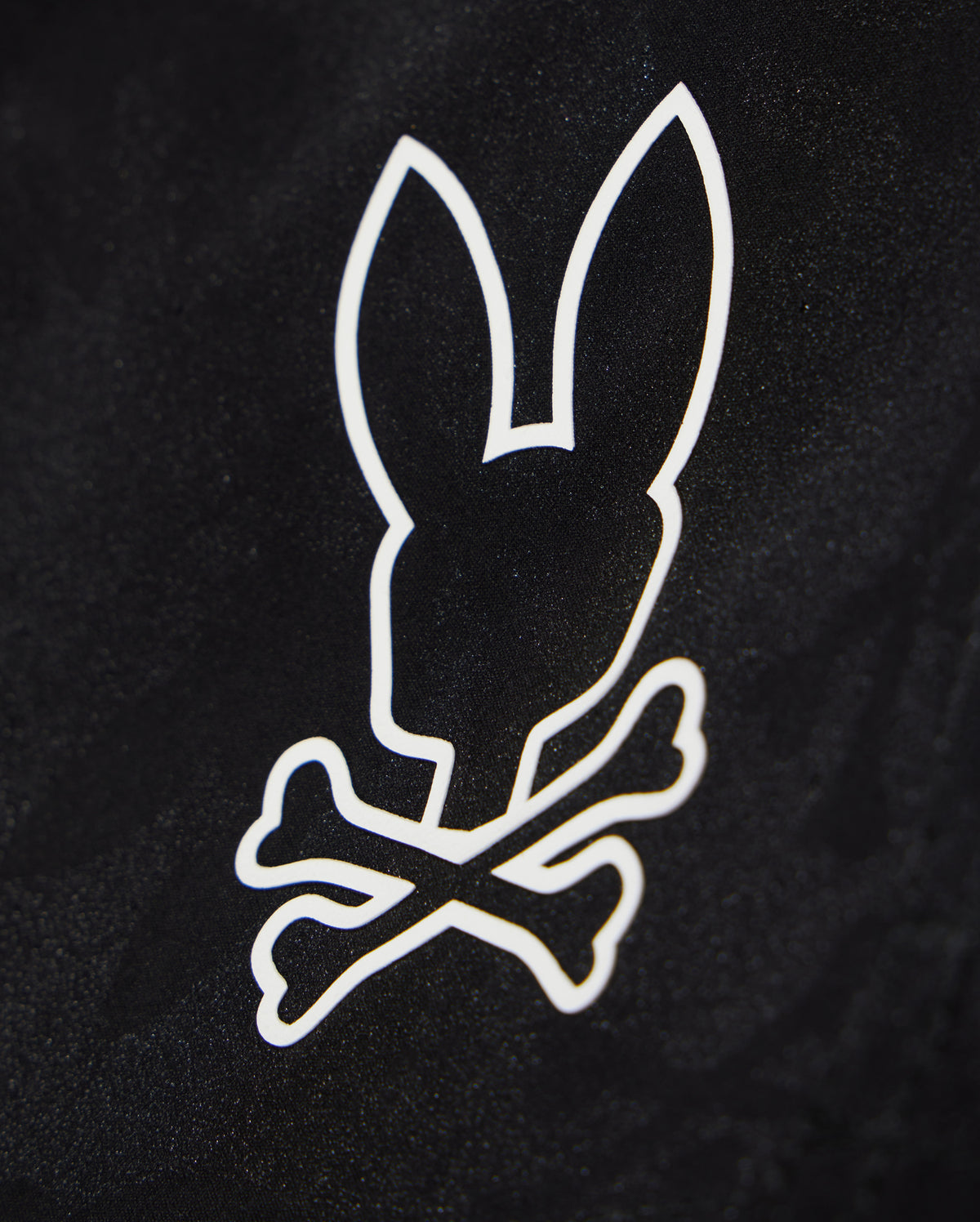 A black background features a white, minimalist graphic of a bunny head with long ears positioned above two crossed bones, resembling a skull and crossbones design on quick-dry material in the KIDS PARKER HYDROCHROMIC SWIM TRUNK - B0W646C200 from Psycho Bunny.