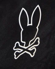 A black background features a white, minimalist graphic of a bunny head with long ears positioned above two crossed bones, resembling a skull and crossbones design on quick-dry material in the KIDS PARKER HYDROCHROMIC SWIM TRUNK - B0W646C200 from Psycho Bunny.