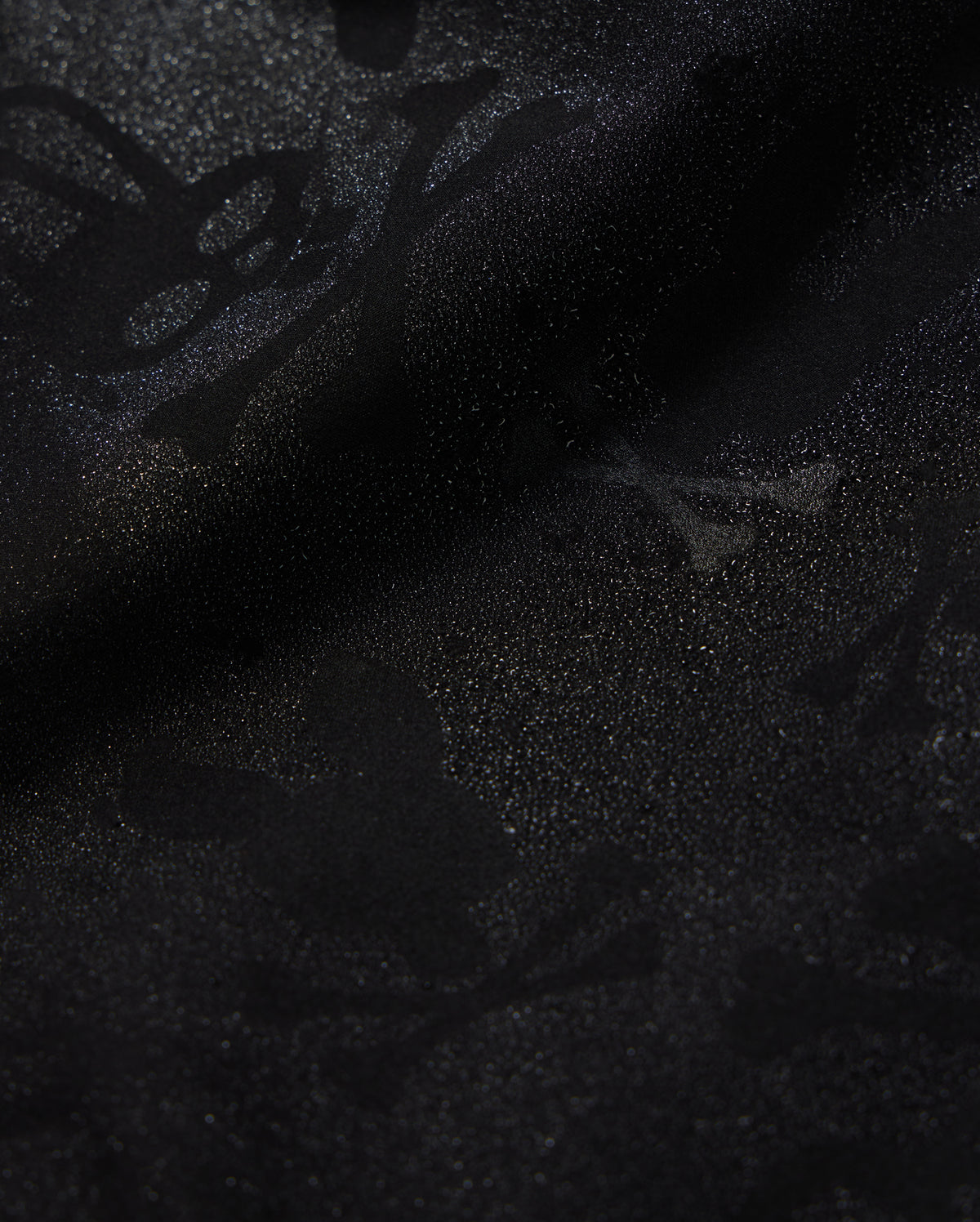 A detailed close-up of a Psycho Bunny KIDS PARKER HYDROCHROMIC SWIM TRUNK - B0W646C200 fabric displaying a subtle textured pattern with skull and crossbones and other abstract shapes. The slightly shiny, quick-dry material adds depth and dimension to the dark hydrochromatic pattern.