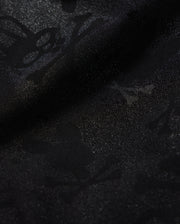 A detailed close-up of a Psycho Bunny KIDS PARKER HYDROCHROMIC SWIM TRUNK - B0W646C200 fabric displaying a subtle textured pattern with skull and crossbones and other abstract shapes. The slightly shiny, quick-dry material adds depth and dimension to the dark hydrochromatic pattern.