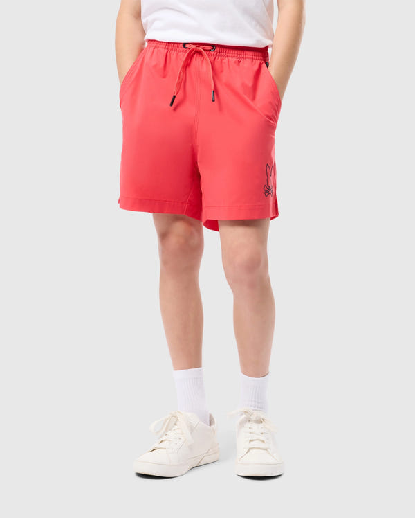 Person wearing a white t-shirt, Psycho Bunny KIDS PARKER HYDROCHROMIC SWIM TRUNK - B0W646C200 with pockets, quick-dry material, white socks, and white sneakers, standing against a gray background. The trunks feature a small black cartoonish bunny face on the lower left side.