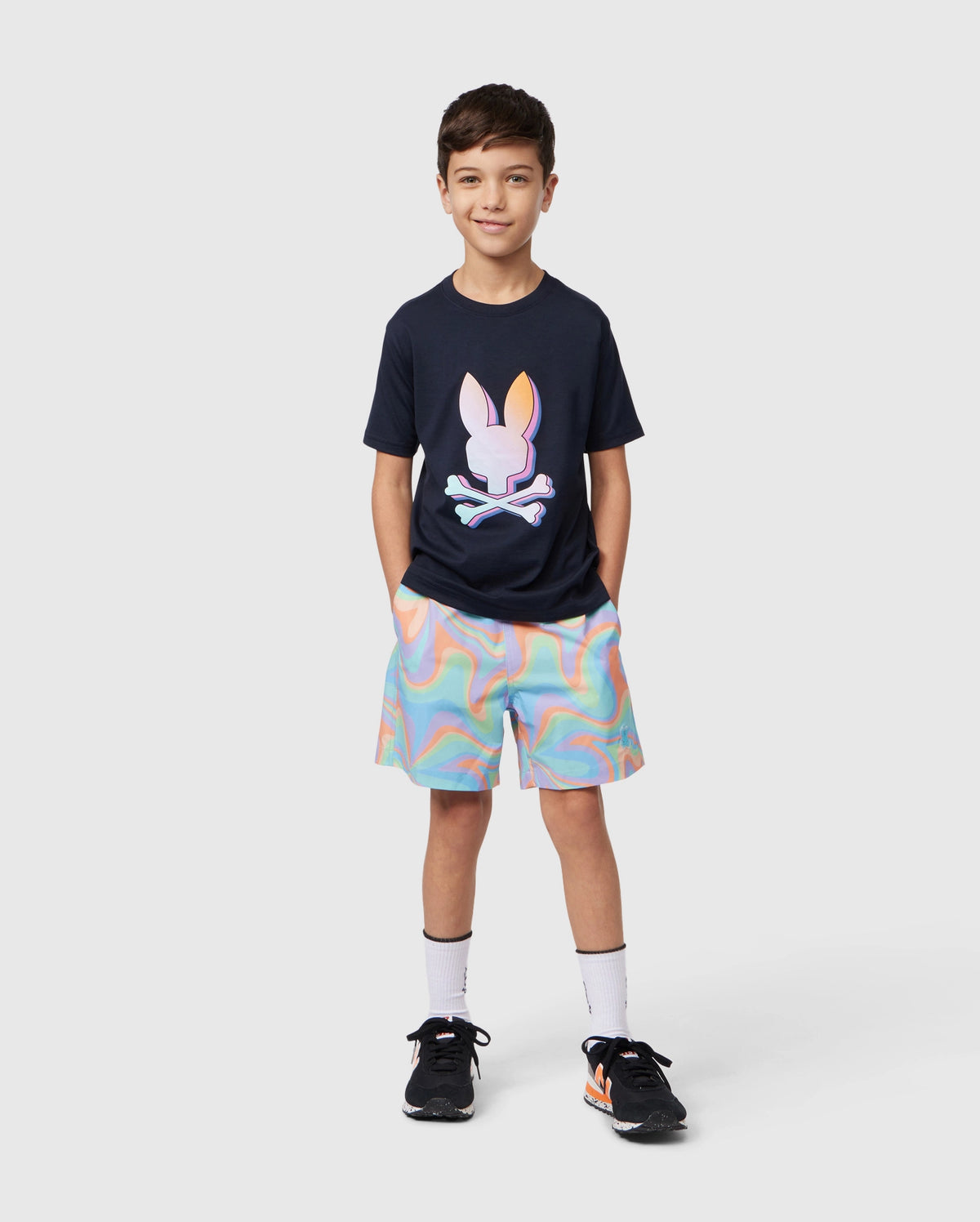 A young boy stands casually with his hands in his pockets, wearing a navy blue t-shirt featuring a colorful bunny skull design. Paired with Psycho Bunny KIDS BLOOMINGTON SWIM TRUNK - B0W941A2PO in a rainbow swirl pattern, he completes the outfit with white socks and black athletic shoes. The background is plain white.
