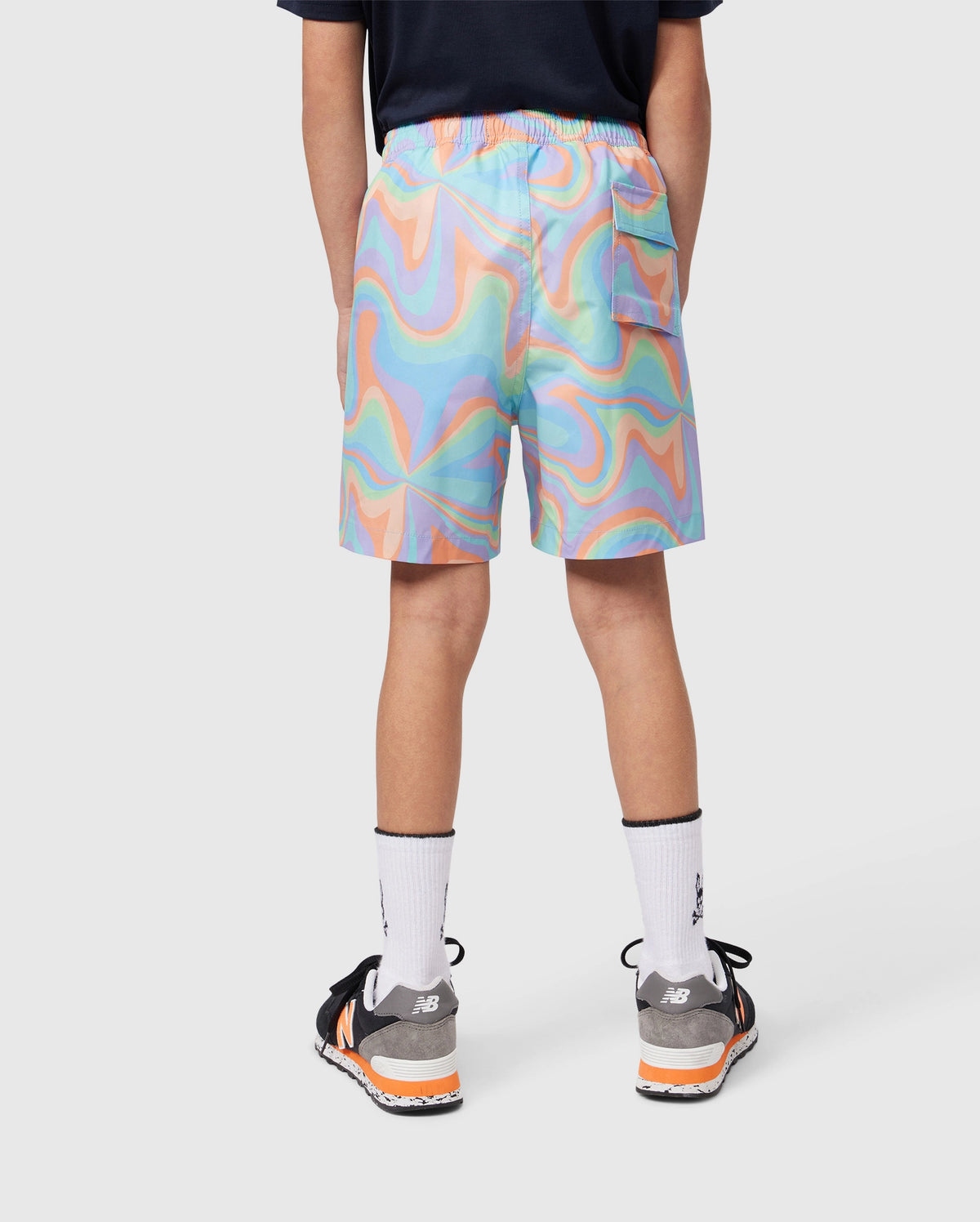 A person wearing colorful, psychedelic-patterned KIDS BLOOMINGTON SWIM TRUNK - B0W941A2PO by Psycho Bunny, white socks, and black and orange sneakers is shown from the back. The person is also wearing a dark t-shirt adorned with a small silicone Bunny patch. The background is plain white.