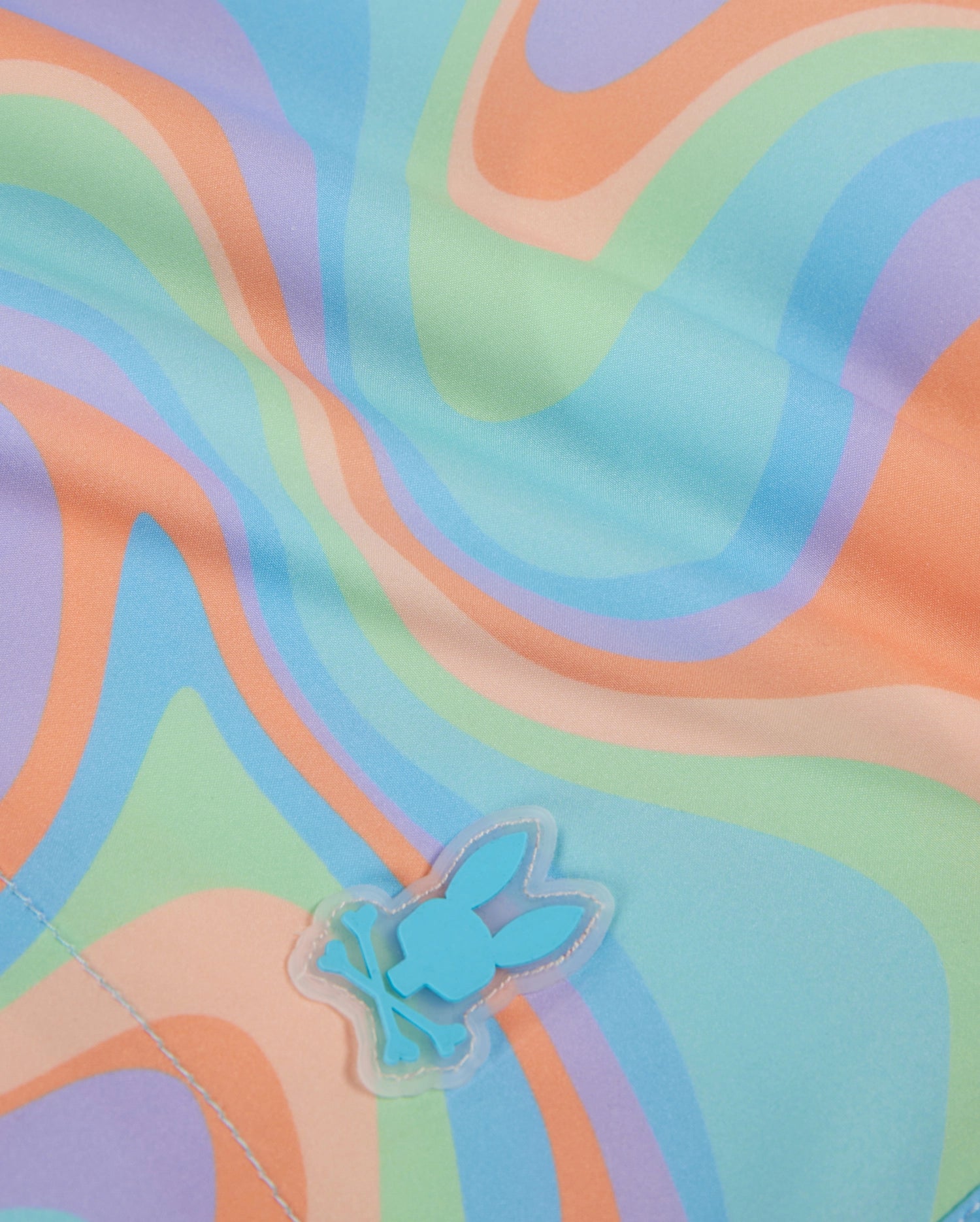 Close-up of a fabric with a colorful, wavy pattern in pastel shades of blue, orange, green, and purple. A small blue silicone Bunny patch featuring a stylized bunny with crossbones is visible near the bottom of the image. There is a visible seam at the bottom left corner. The product shown is the KIDS BLOOMINGTON SWIM TRUNK - B0W941A2PO by Psycho Bunny.