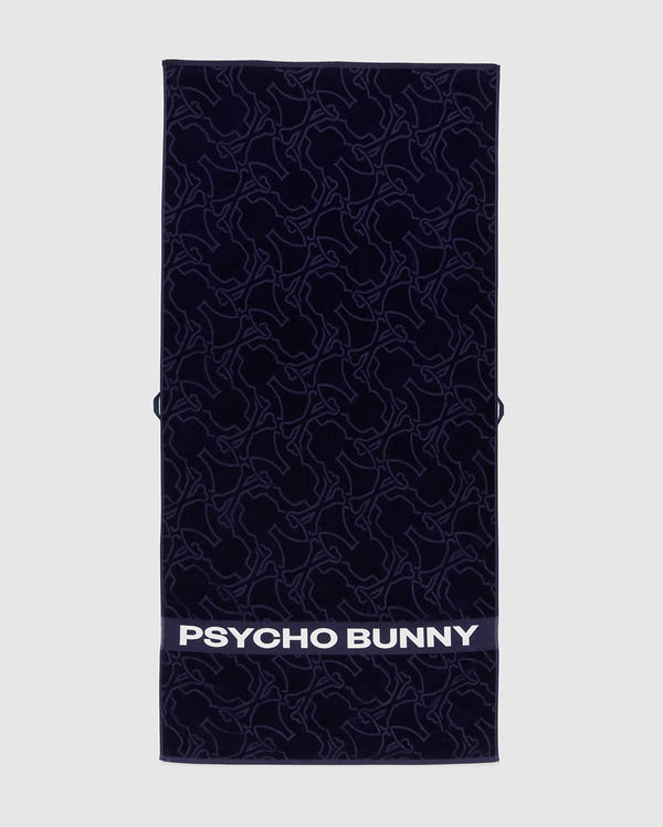 The Towel - B6A287F200 by Psycho Bunny is a rectangular dark towel made from premium terrycloth cotton, featuring an intricate bunny pattern. The bottom displays "PSYCHO BUNNY" in bold white letters, showcasing high-quality design.