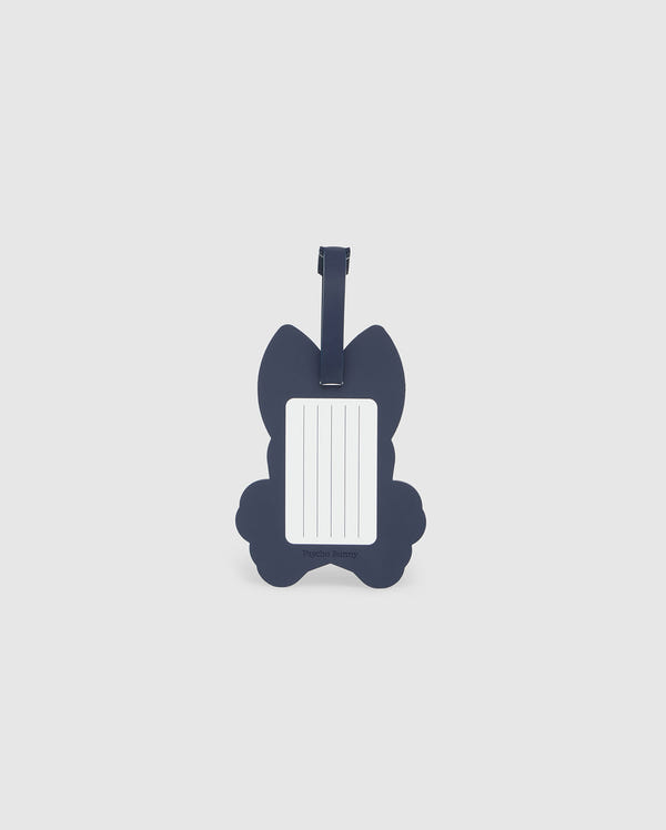 A navy blue luggage tag in the shape of a cat is pictured. The front side features a rectangular white label with blank lines for writing contact information. Made from detachable silicone, the Psycho Bunny MENS LUGGAGE TAG - B6A387B200 has a strap at the top for attaching to luggage.