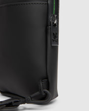 Close-up of the Psycho Bunny MENS RUBBERIZED CROSSBODY - B6A635F200, featuring sleek black leather with a visible zipper and a small rabbit-shaped metal charm. The durable design includes a side clip attachment and hints of green lining peeking by the zipper.