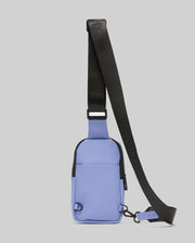 The Psycho Bunny MENS RUBBERIZED CROSSBODY - B6A635F200 is a compact, rectangular light purple bag in durable polyester with a wide, adjustable black strap and zipper, shown upright on a plain light gray background.