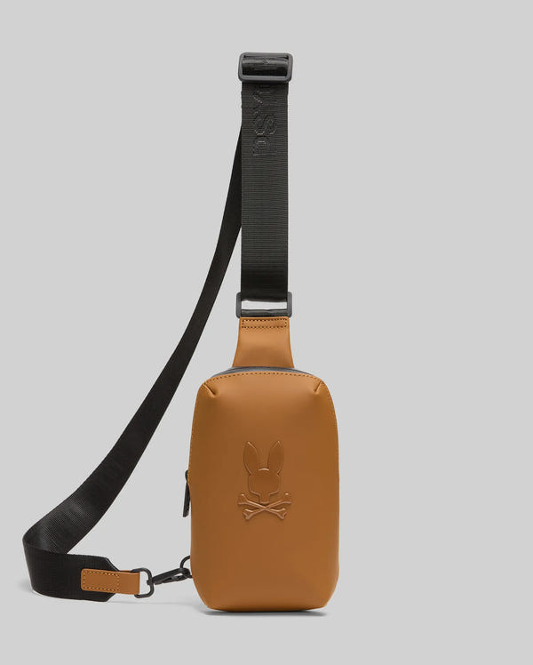The Psycho Bunny MENS RUBBERIZED CROSSBODY - B6A635F200 is a tan leather bag with a black adjustable strap and embossed bunny and crossbones logo on the front, shown against a plain light grey background.