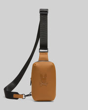 The Psycho Bunny MENS RUBBERIZED CROSSBODY - B6A635F200 is a tan leather bag with a black adjustable strap and embossed bunny and crossbones logo on the front, shown against a plain light grey background.