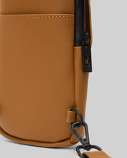 Close-up of the Psycho Bunny MENS RUBBERIZED CROSSBODY - B6A635F200 in tan polyester with a black zipper, small bunny logo, black hardware, and an adjustable detachable strap against a light gray background.