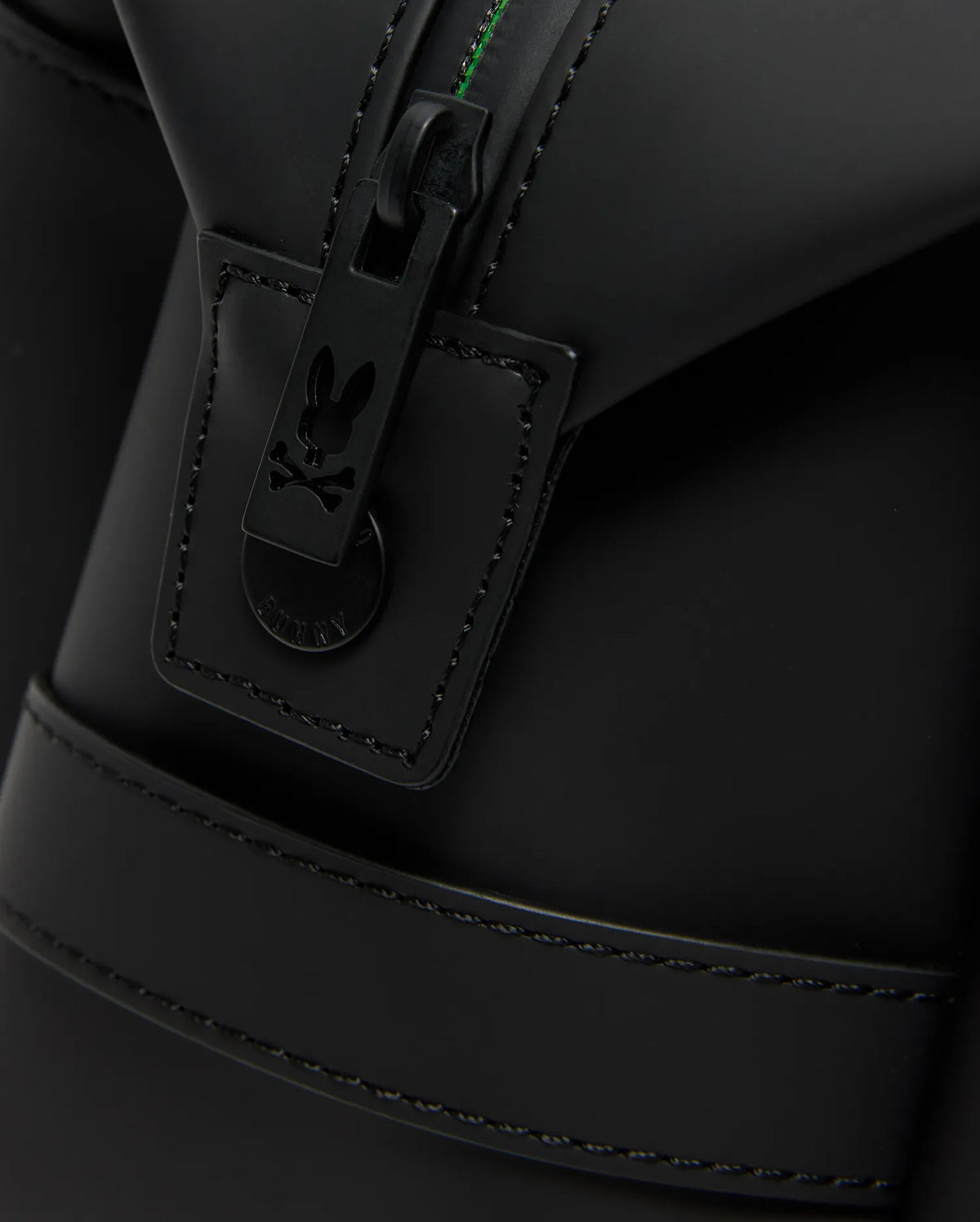 Close-up of the Psycho Bunny MENS RUBBERIZED TOILETRY KIT (B6A636F200) shows a black matte zip with a bunny skull and crossbones pull tab, detailed stitching, and green thread—ideal for keeping your essentials secure.