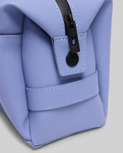 Close-up of the Psycho Bunny MENS RUBBERIZED TOILETRY KIT (B6A636F200) in light purple, made of smooth material with a black zip pouch, black button, visible stitching, set against a grey background.