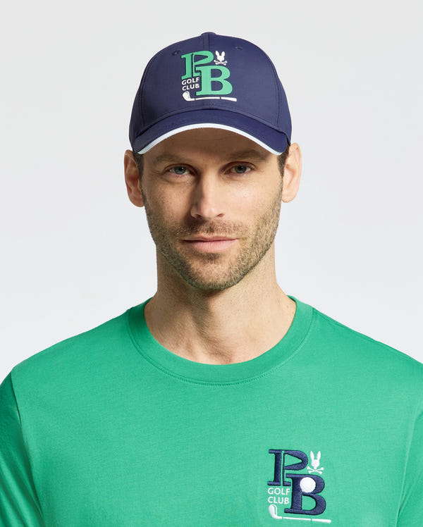 A man in a green cotton-blend top with a "PB Golf Club" logo, paired with the Psycho Bunny MENS DENIS SPORT CAP in navy, featuring the same logo and rabbit emblem. Both items are sporty and practical for an active day on the course.