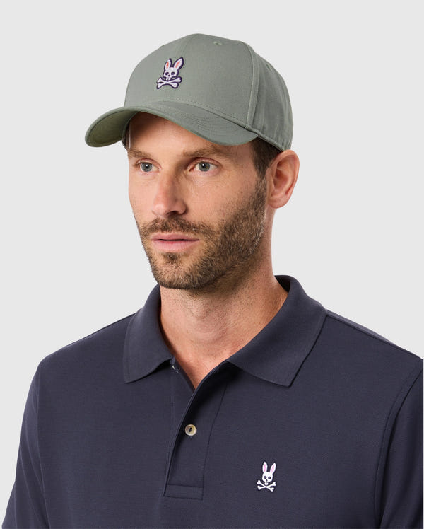 A bearded man wears a Psycho Bunny 100% cotton classic men's baseball cap (product B6A816B200) in a solid pattern and navy polo shirt, both featuring a rabbit with crossed bones logo, against a plain background.