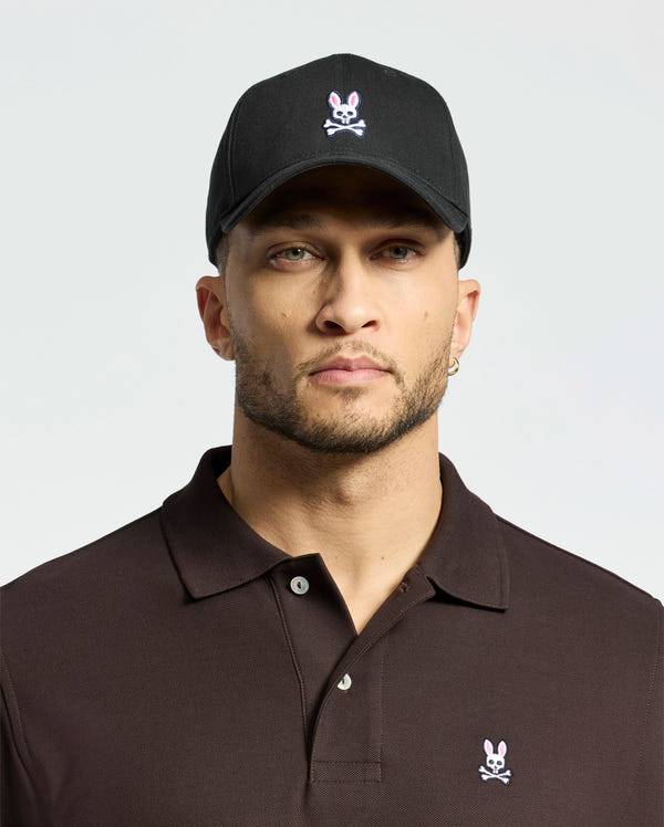 A man wears a MENS CLASSIC BASEBALL CAP - B6A816A1HT by Psycho Bunny, featuring the iconic embroidered Bunny logo. The black cap complements his brown polo shirt, trimmed beard, earring, and laid-back style with its adjustable fit and neutral expression.