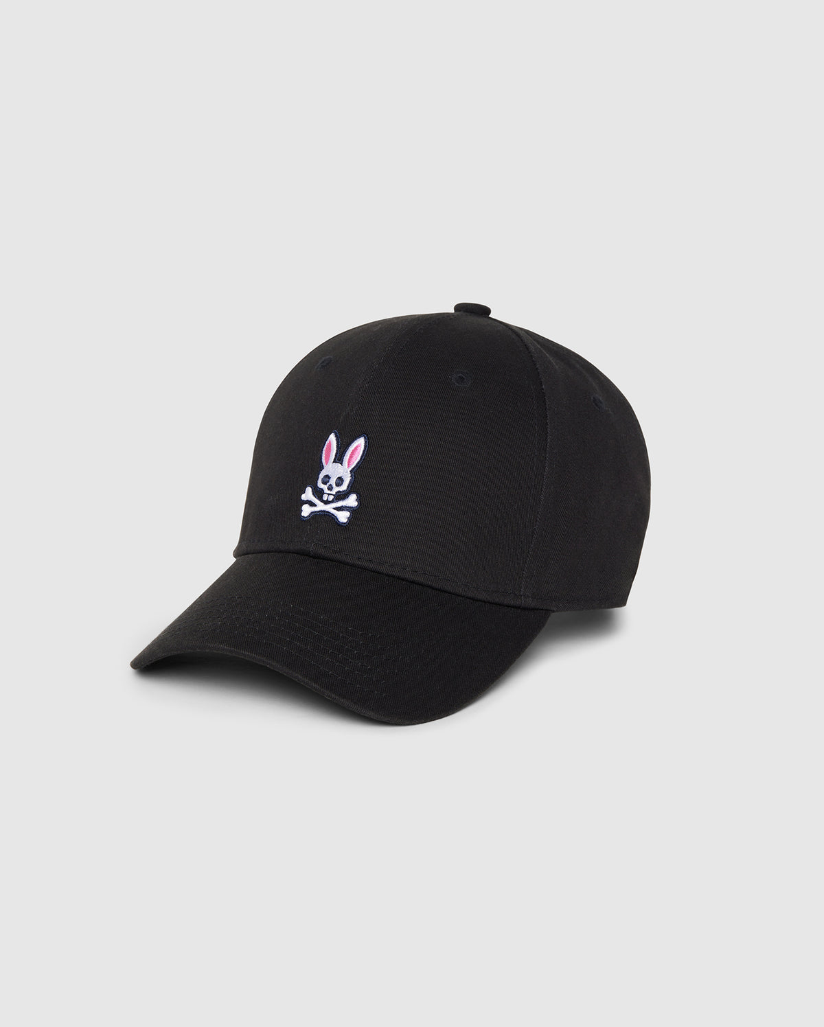 A Psycho Bunny MENS CLASSIC BASEBALL CAP - B6A816A1HT in black featuring a small embroidered pink and white bunny above crossed bones on the front. Made from durable cotton twill, it has a curved brim and six panels with eyelets for ventilation. The background is plain and light gray.
