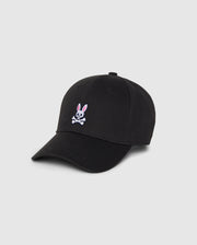 A Psycho Bunny MENS CLASSIC BASEBALL CAP - B6A816A1HT in black featuring a small embroidered pink and white bunny above crossed bones on the front. Made from durable cotton twill, it has a curved brim and six panels with eyelets for ventilation. The background is plain and light gray.
