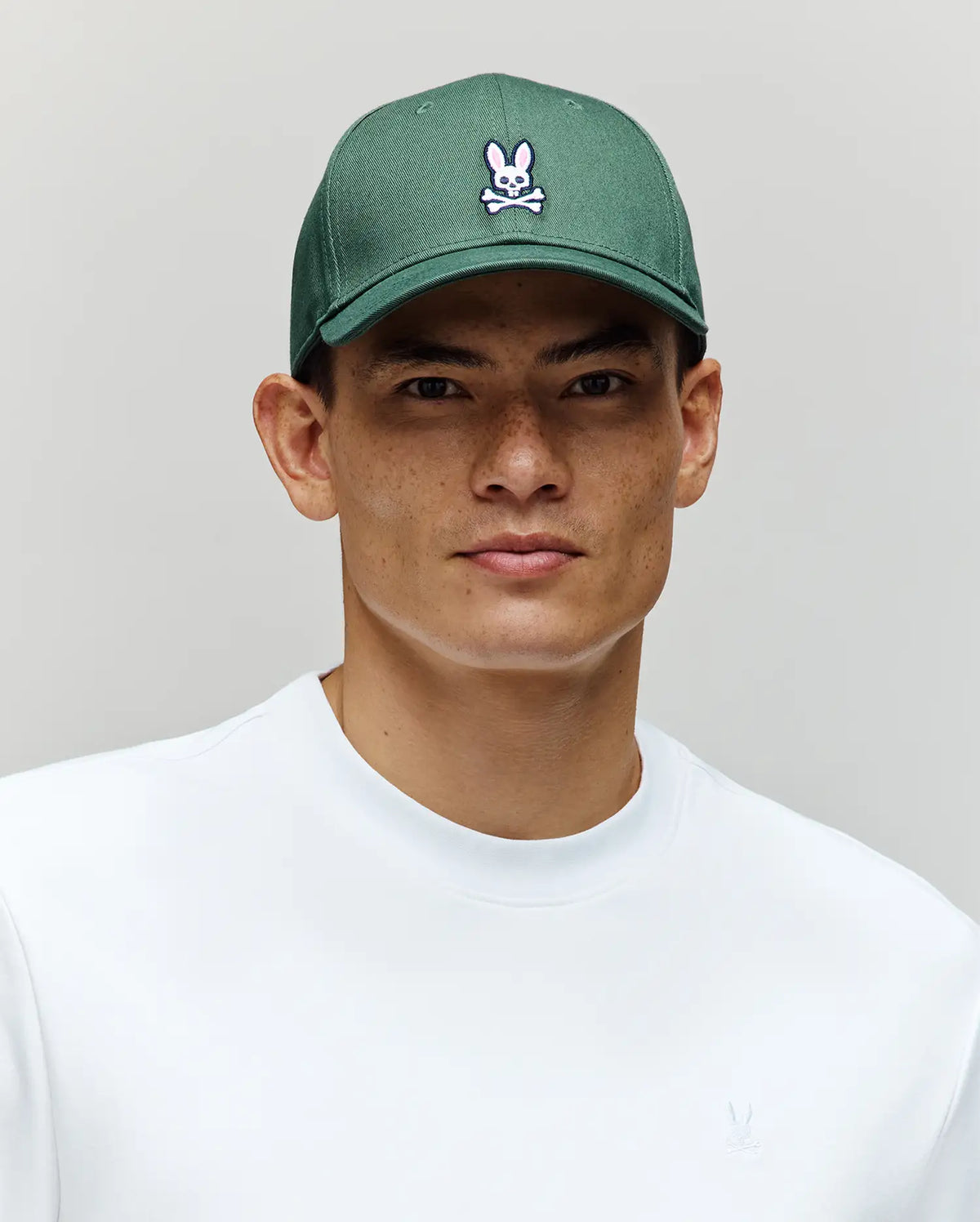 A young man wears a white shirt and the Psycho Bunny MENS CLASSIC BASEBALL CAP (B6A816A1HT)—a green cotton hat with a curved brim and embroidered Bunny logo—against a plain light gray background.
