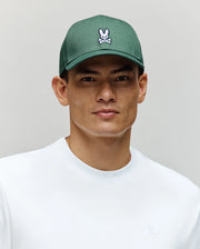 A young man wears a white shirt and the Psycho Bunny MENS CLASSIC BASEBALL CAP (B6A816A1HT)—a green cotton hat with a curved brim and embroidered Bunny logo—against a plain light gray background.