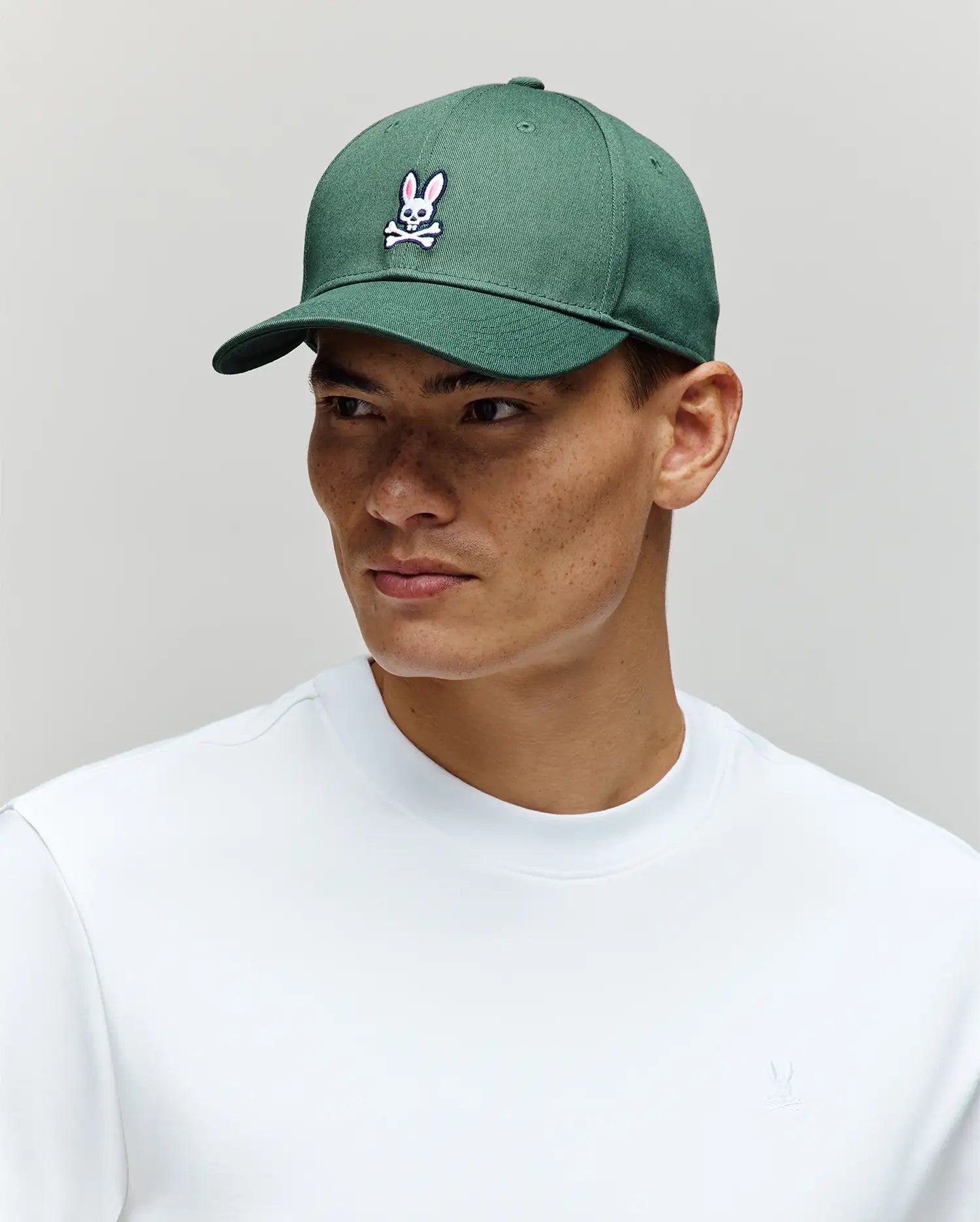 A person wears the Psycho Bunny MENS CLASSIC BASEBALL CAP - B6A816A1HT, an adjustable dark green cotton hat with a curved brim and embroidered Bunny logo, paired with a white crew neck shirt, looking slightly to the side against a neutral background.