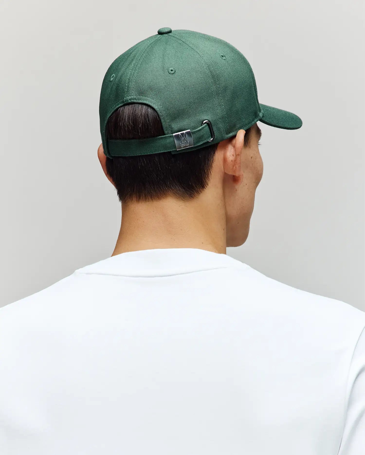 A person is shown from behind wearing a Psycho Bunny MENS CLASSIC BASEBALL CAP - B6A816A1HT with an adjustable strap and curved brim, along with a plain white shirt, set against a neutral background.