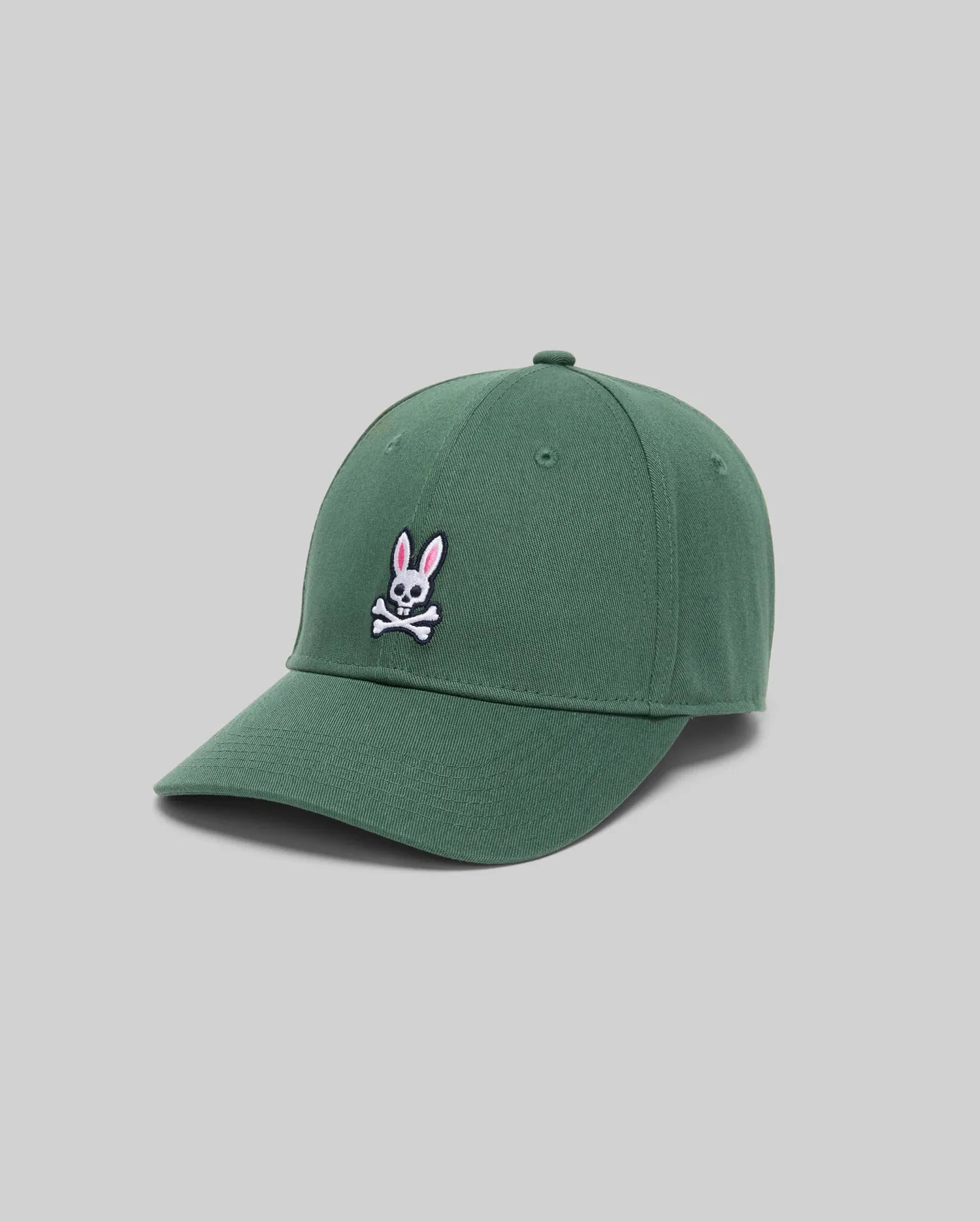 The Psycho Bunny MENS CLASSIC BASEBALL CAP (B6A816A1HT) is a green cotton cap with a white skull and crossbones and pink bunny ears, adjustable fit, curved brim, shown against a plain light gray background.