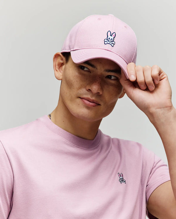 A young man wearing a light pink T-shirt and the Psycho Bunny MENS CLASSIC BASEBALL CAP (B6A816A1HT) tilts its adjustable brim, looking off to the side against a plain background.