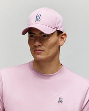 A young man wears the Psycho Bunny MENS CLASSIC BASEBALL CAP - B6A816A1HT and a matching pink shirt, both featuring the signature Bunny and crossbones logo, standing against a plain light gray background. The cap offers an adjustable fit for comfort.