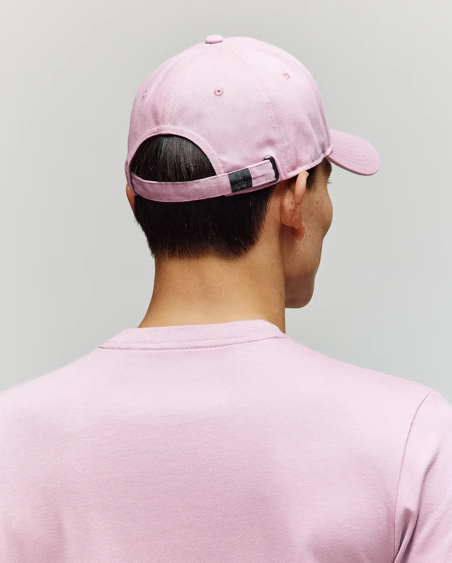 A person wearing a Psycho Bunny MENS CLASSIC BASEBALL CAP - B6A816A1HT in light pink with an embroidered Bunny and a matching pink t-shirt is shown from behind against a plain light gray background.