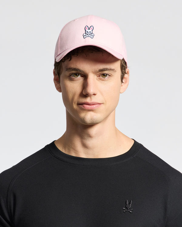 A person wears a Psycho Bunny MENS CLASSIC BASEBALL CAP - B6A816A1HT, featuring a pink classic design with a cartoon skull and crossbones with bunny ears. Made from soft cotton twill for an adjustable fit, this look is completed with a black shirt on a plain white backdrop.