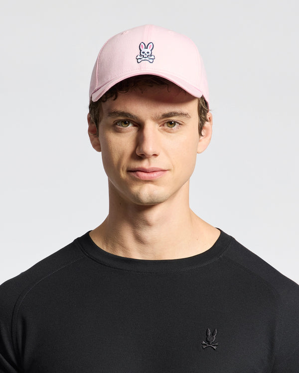 A person wears a Psycho Bunny MENS CLASSIC BASEBALL CAP - B6A816A1HT, featuring a pink classic design with a cartoon skull and crossbones with bunny ears. Made from soft cotton twill for an adjustable fit, this look is completed with a black shirt on a plain white backdrop.