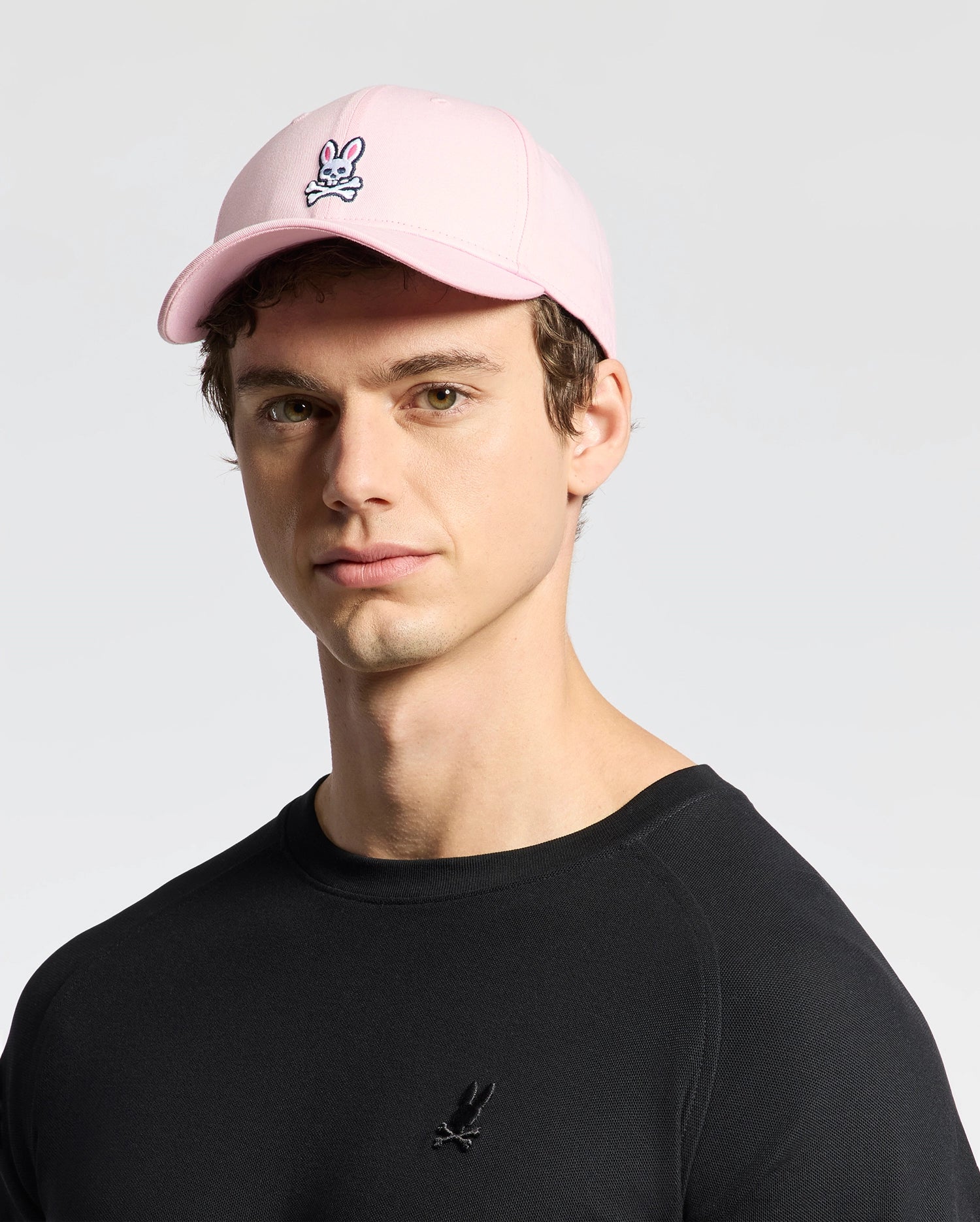 A man models Psycho Bunny's MENS CLASSIC BASEBALL CAP (B6A816A1HT), a light pink cotton twill cap with an adjustable fit featuring the bunny logo, paired with a black shirt against a plain background.