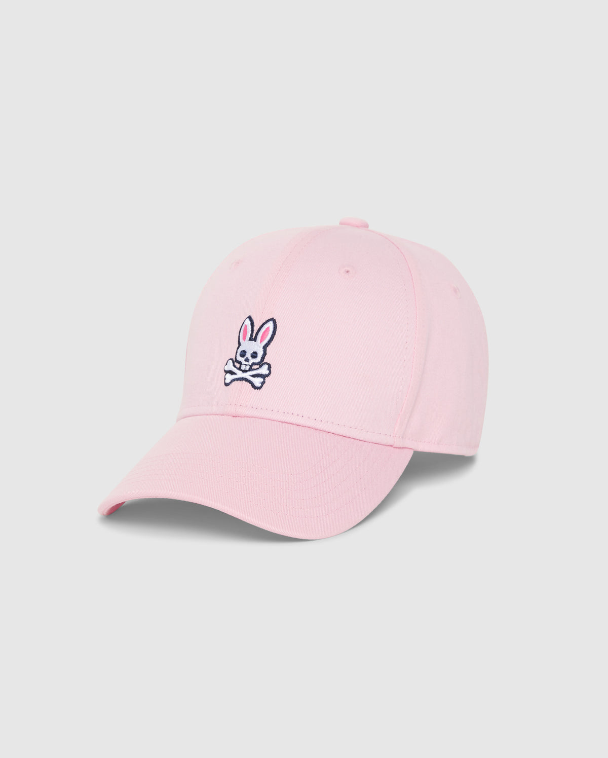 The MENS CLASSIC BASEBALL CAP - B6A816A1HT by Psycho Bunny is a timeless light pink baseball cap, meticulously crafted from 100% cotton, and showcased against a plain background. It features an embroidered design on the front, depicting a white bunny head with crossbones underneath. The bunny sports pink ears and black outlines, imparting a playful touch to this classic accessory.
