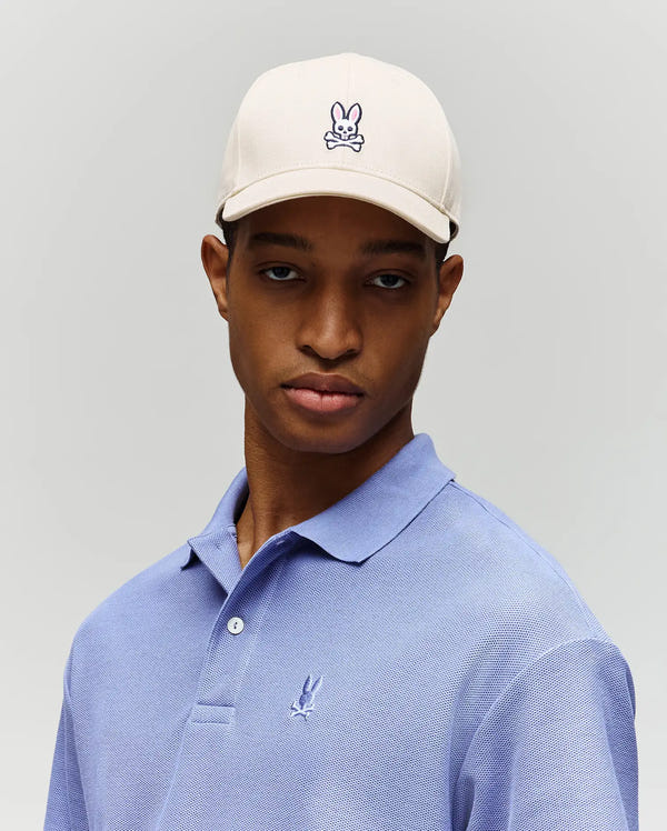 A young man wearing the Psycho Bunny MENS CLASSIC BASEBALL CAP (B6A816A1HT), a cream-colored cotton cap with an embroidered bunny skull logo and adjustable closure, and a light purple polo with a small bird logo stands against a plain light gray background.