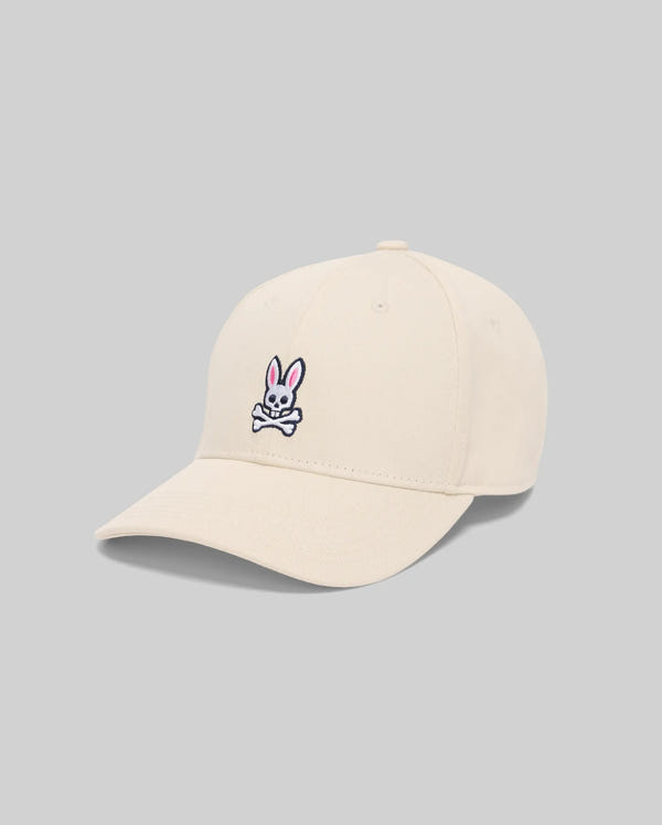 The Psycho Bunny MENS CLASSIC BASEBALL CAP - B6A816A1HT is a beige adjustable cotton cap with an embroidered bunny skull and crossbones with pink ears on the front, shown against a plain gray background.