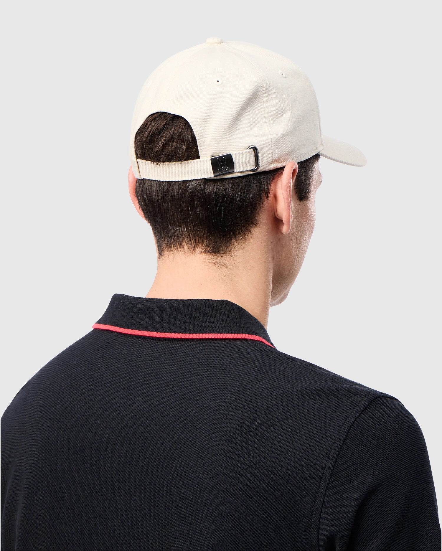 A person wearing the Psycho Bunny MENS CLASSIC BASEBALL CAP - B6A816B200, crafted from 100% cotton in classic beige with an adjustable strap, is seen from behind. They are dressed in a black shirt featuring red piping on the collar against a plain gray background.