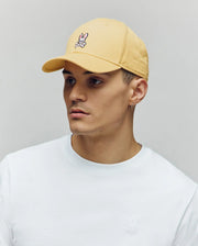 A young man in a plain white tee and a Psycho Bunny MENS CLASSIC BASEBALL CAP (B6A816F200) with bunny logo and adjustable fit, plus a small hoop earring, looks to the side against a neutral background.