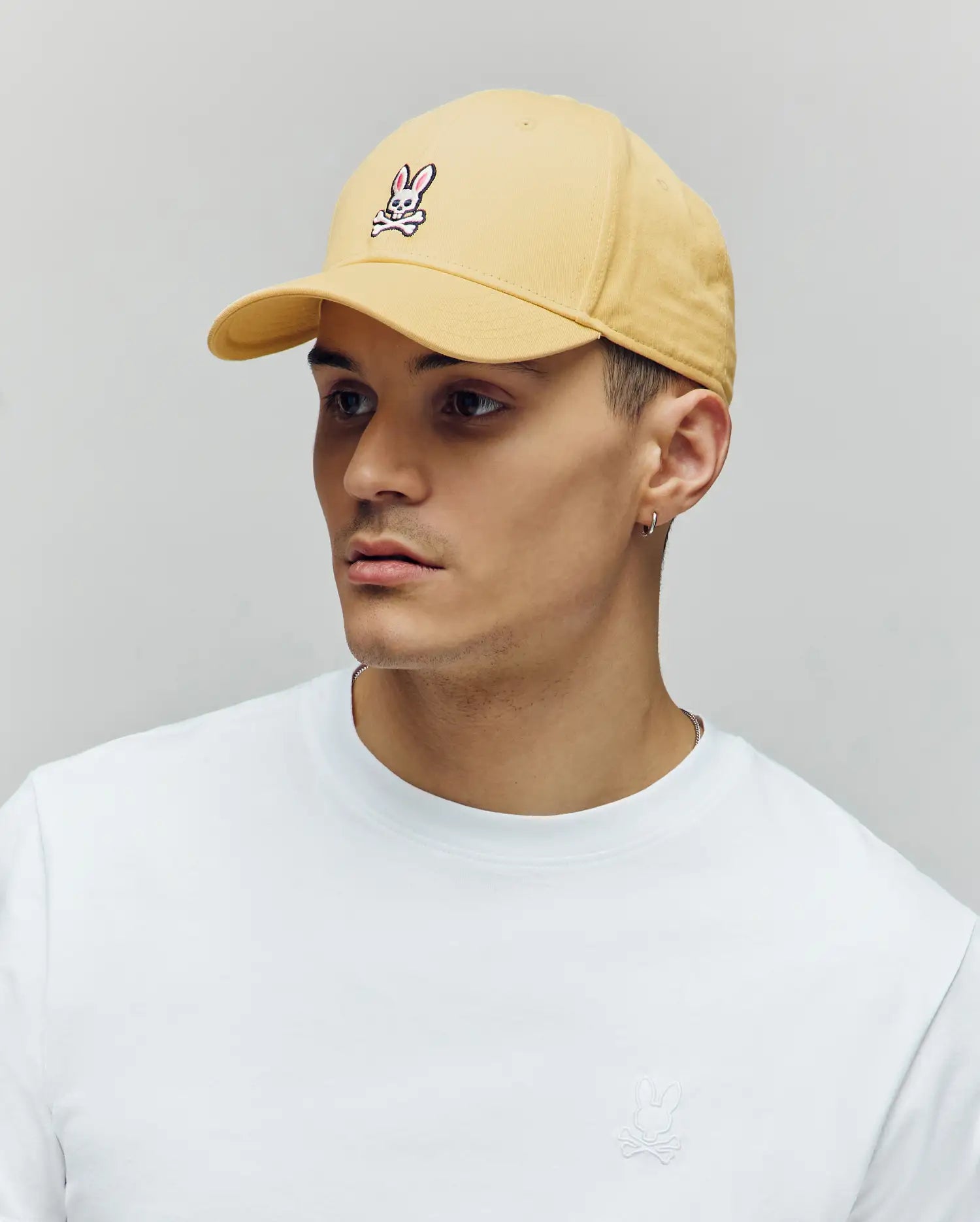 A young man in a plain white tee and a Psycho Bunny MENS CLASSIC BASEBALL CAP (B6A816F200) with bunny logo and adjustable fit, plus a small hoop earring, looks to the side against a neutral background.