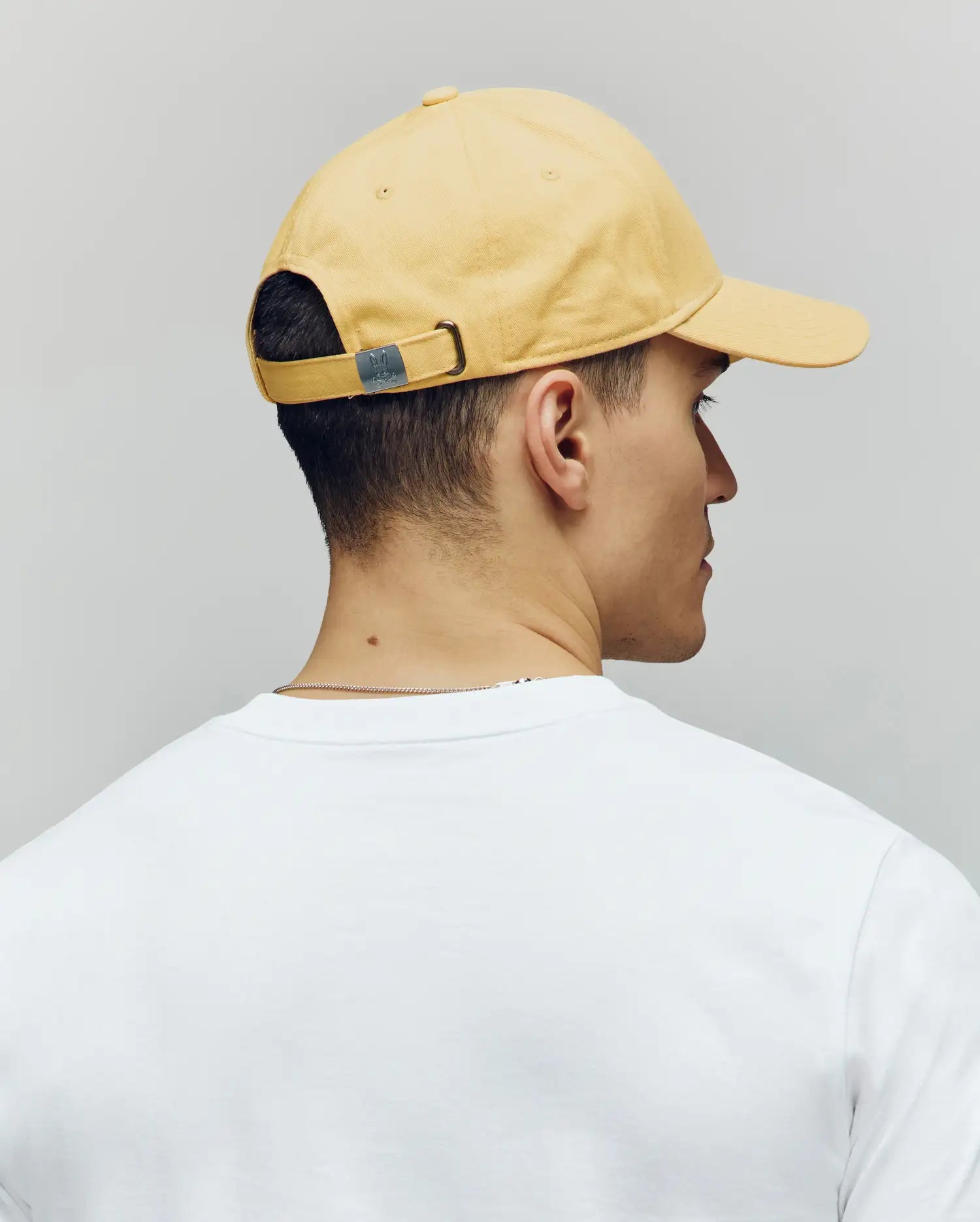 A person is seen from behind, facing right, wearing a yellow Psycho Bunny MENS CLASSIC BASEBALL CAP - B6A816F200 with an adjustable fit and a white shirt against a plain gray background.