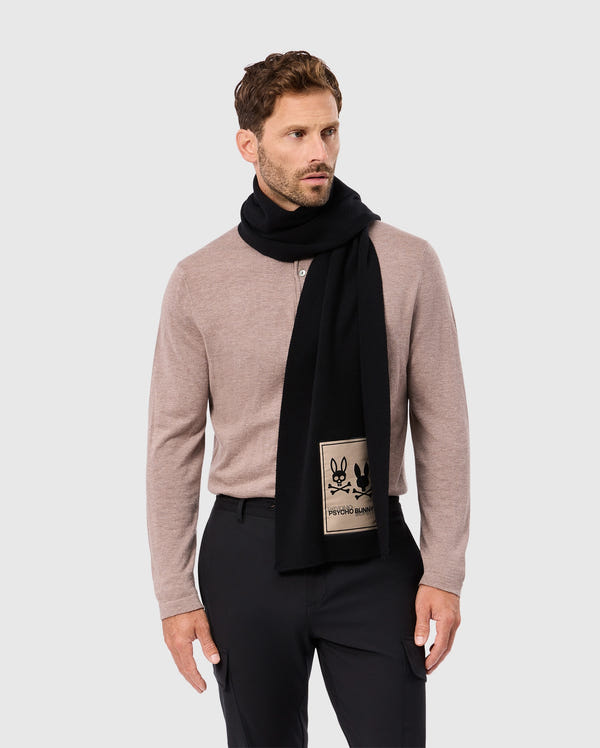 A man wearing a beige long-sleeve shirt stands against a gray background, elegantly adorned with Psycho Bunny's luxury MENS MONROE TWILL PATCH SCARF (B6A892D200) crafted from a black cotton wool blend. The scarf is distinguished by a charming bunny design patch. He has short dark hair and a beard, looking to his left.