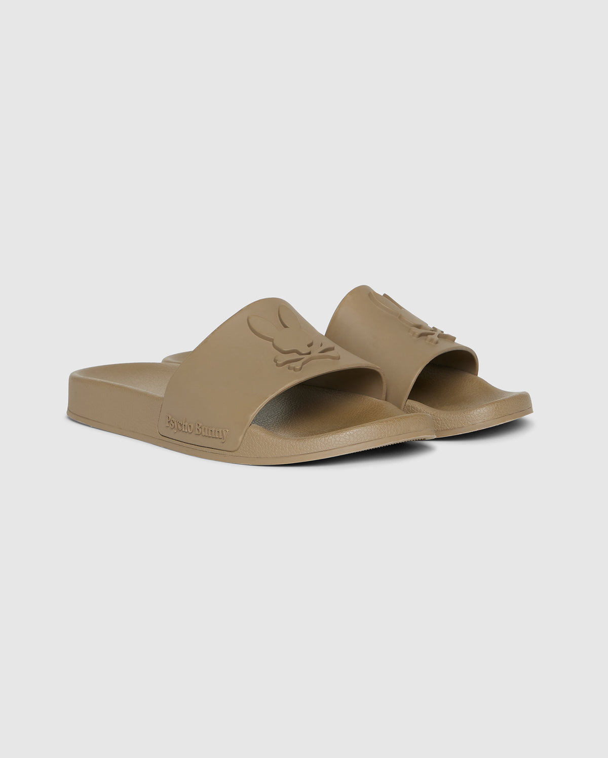 A pair of beige Psycho Bunny pool slides with molded footbeds and embossed logo on the straps, isolated on a white background.