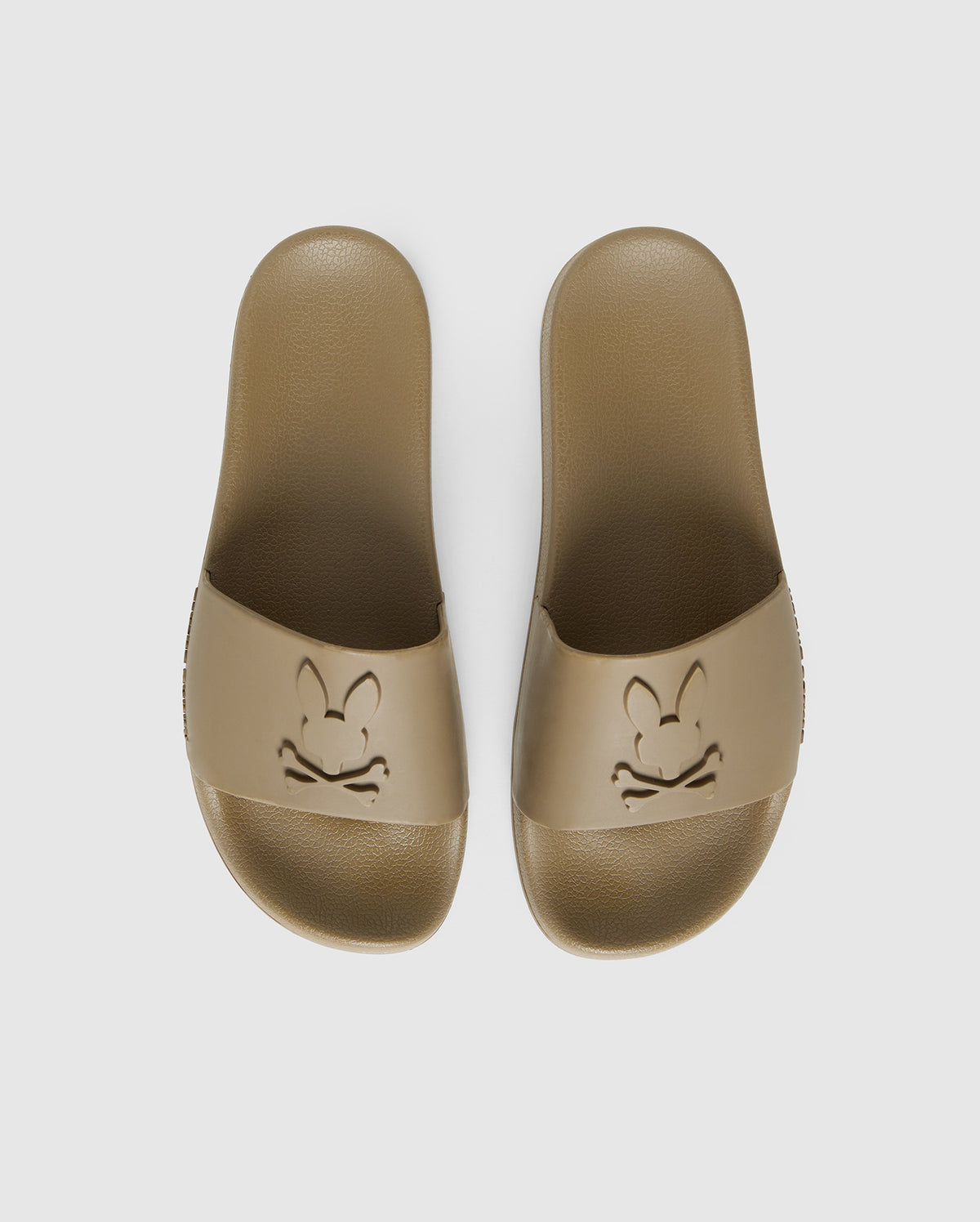 A pair of brown pool slides with an embossed cartoon rabbit face design on each top strap, viewed from above on a white background are the MENS LOGO SLIDES - B6A910U1FT by Psycho Bunny.