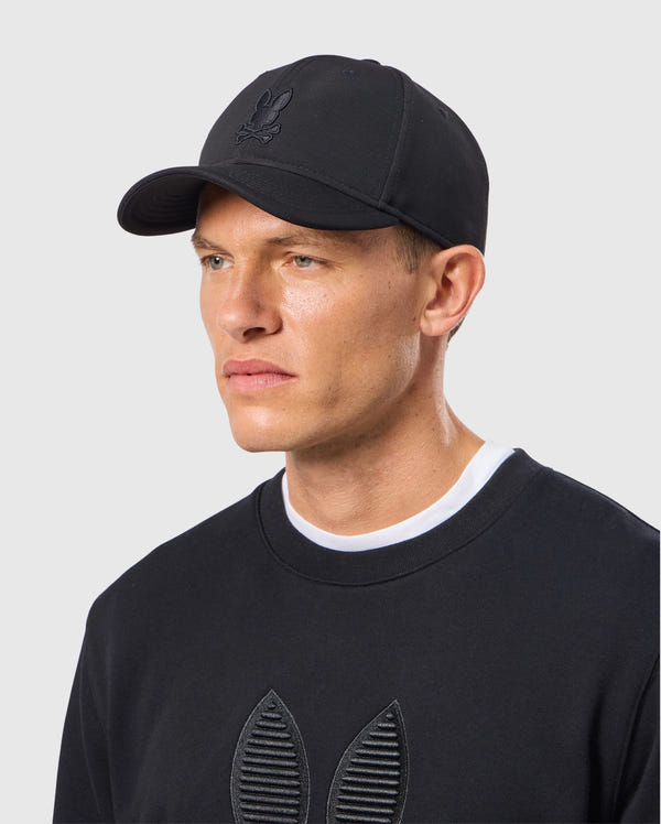 A person wearing a black MENS JAMES PREMIUM BASEBALL CAP - B6A953D200 by Psycho Bunny and a matching black sweatshirt featuring an embroidered Bunny logo, set against a plain gray background.
