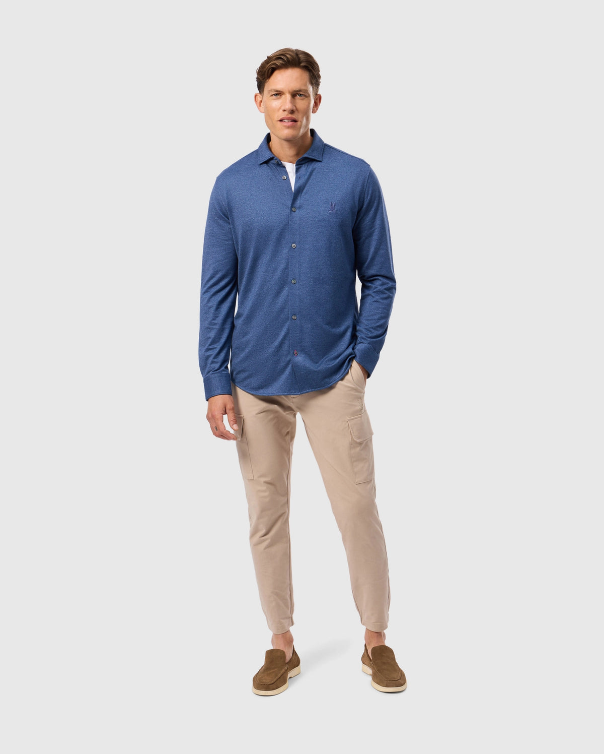A man in a blue Psycho Bunny MENS DALTON KNIT SHIRT - B6C747D200 and beige pants stands against a light background. Brown slip-on shoes complete his look as his hands rest at his sides, showcasing an effortlessly stylish everyday outfit.