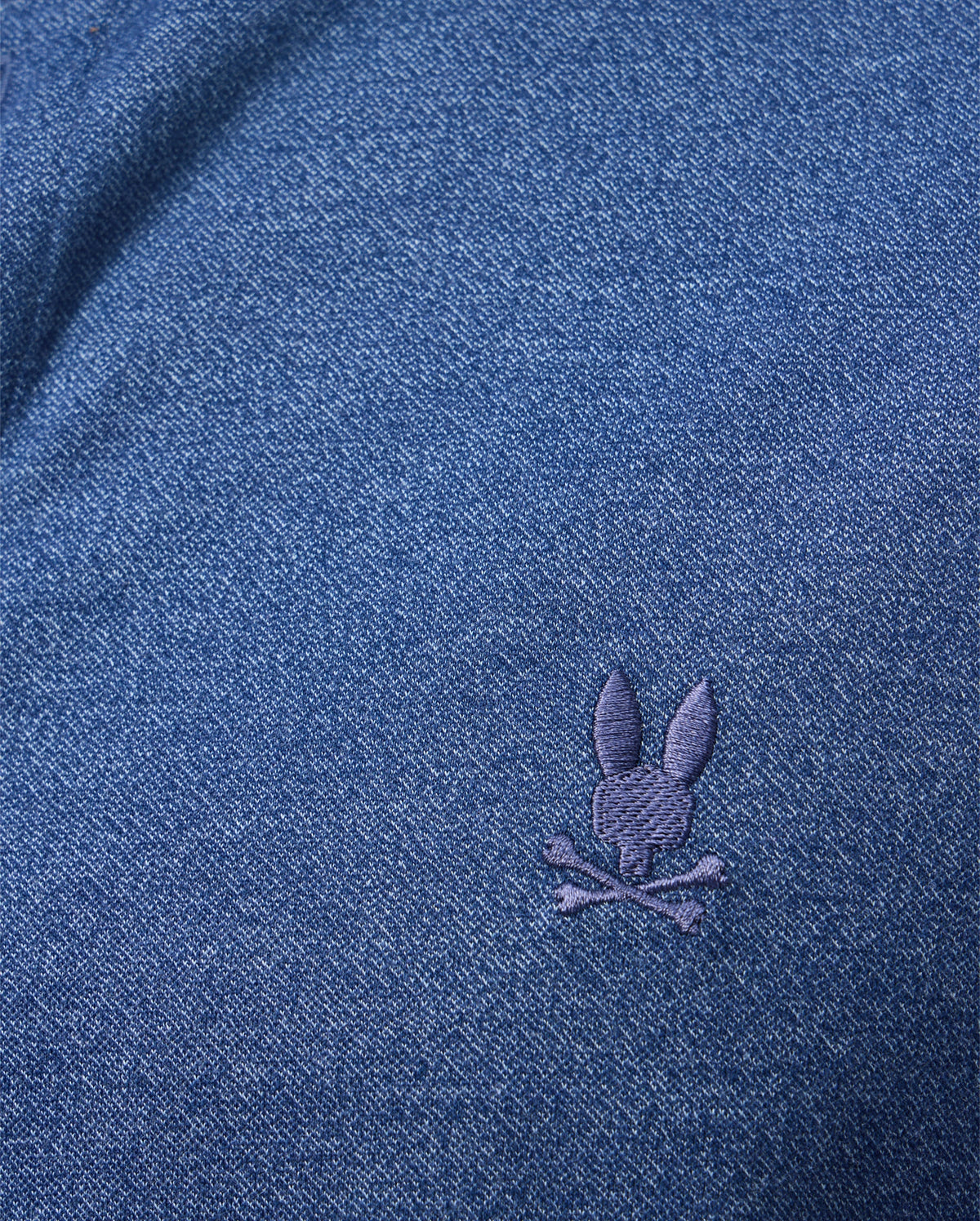 A detailed view of the MENS DALTON KNIT SHIRT by Psycho Bunny showcases a dark blue fabric with a subtle texture and an embroidered bunny head above crossed bones in lighter blue thread. Made from luxurious Peruvian Pima cotton, this shirt offers exceptional softness and comfort.